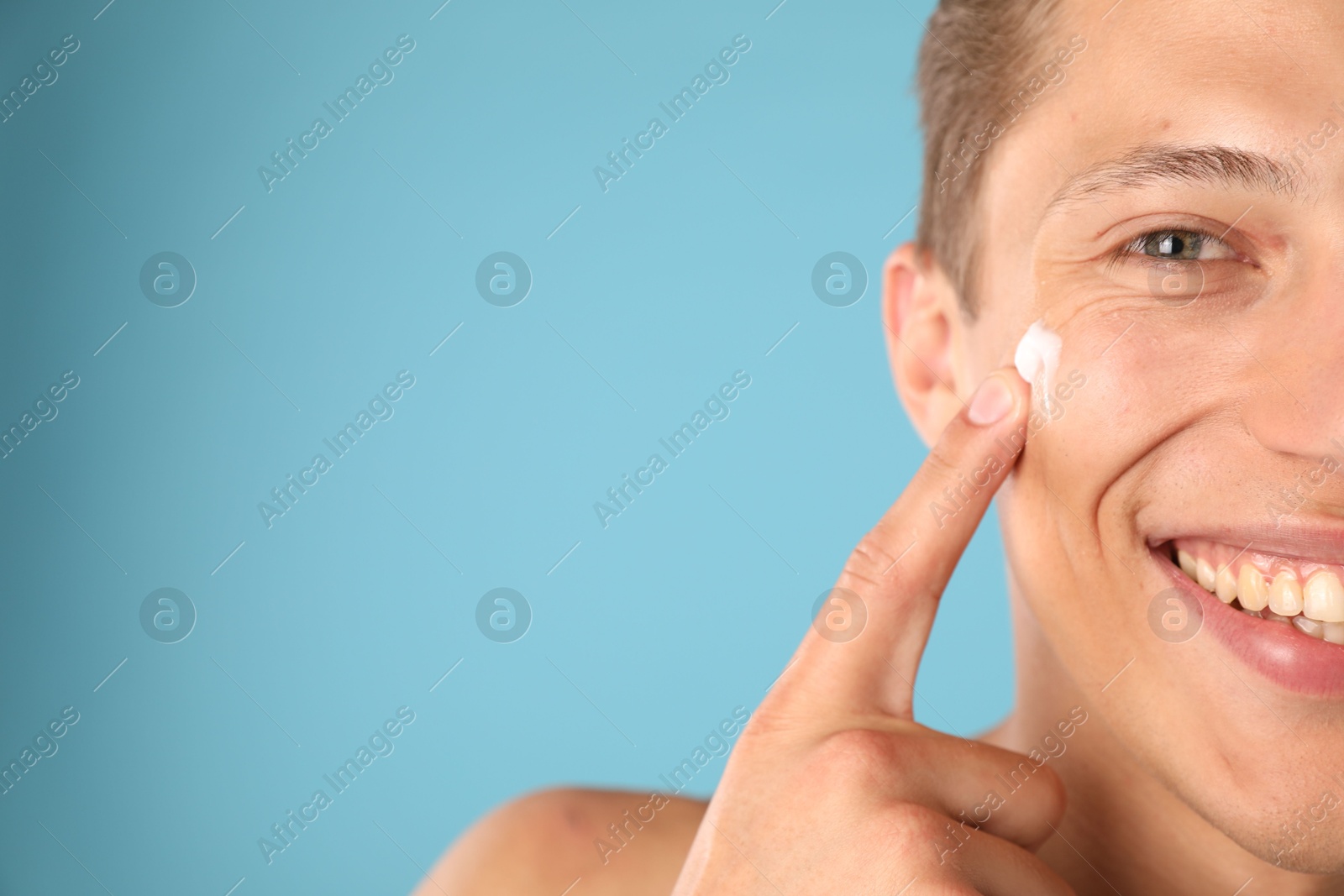 Photo of Handsome young man applying cream onto his face on light blue background, closeup. Space for text