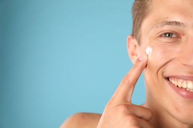 Photo of Handsome young man applying cream onto his face on light blue background, closeup. Space for text