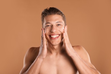 Photo of Handsome young man applying cream onto his face on light brown background