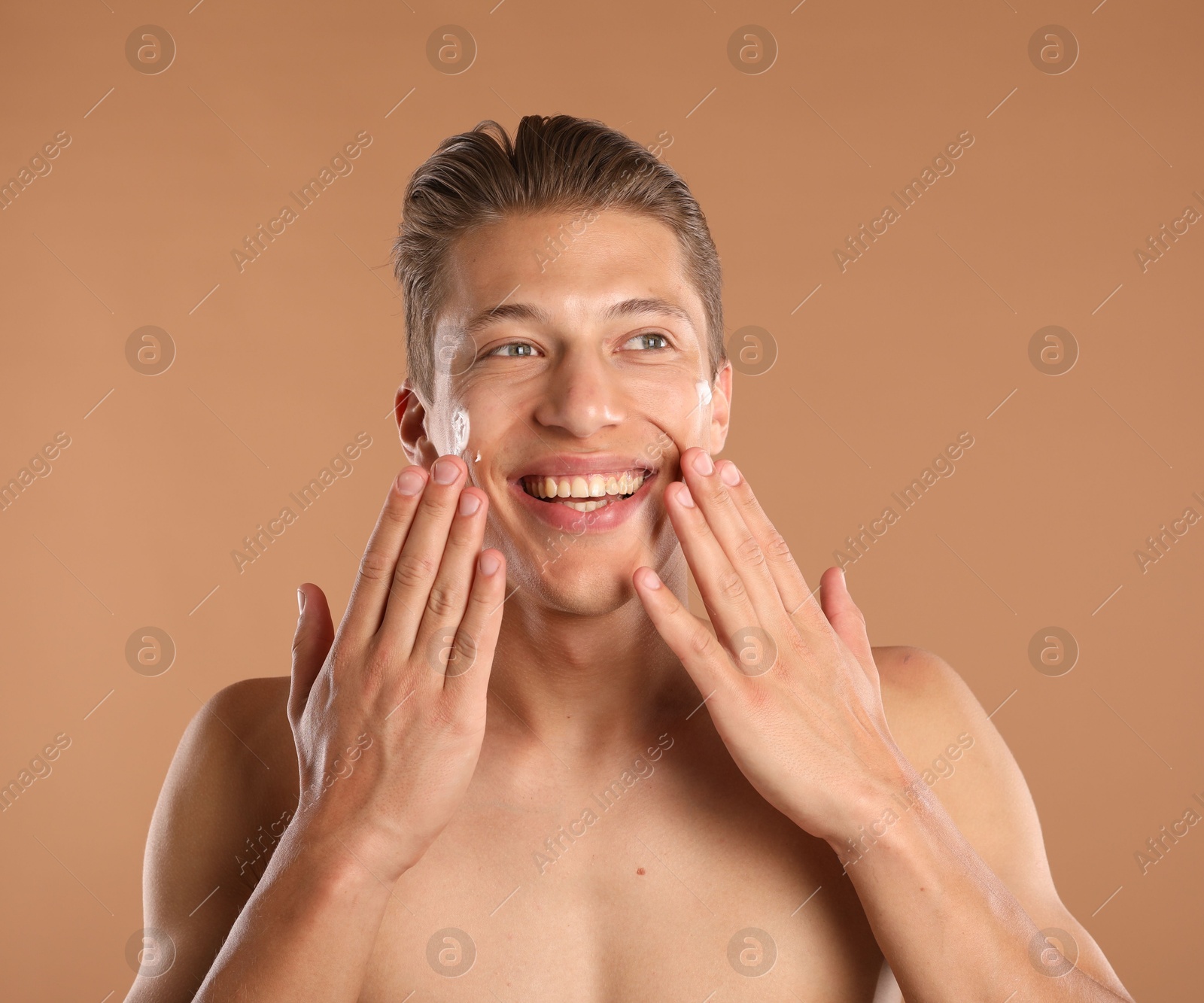 Photo of Handsome young man applying cream onto his face on light brown background
