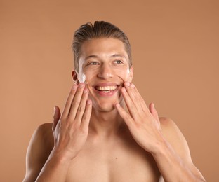 Photo of Handsome young man applying cream onto his face on light brown background
