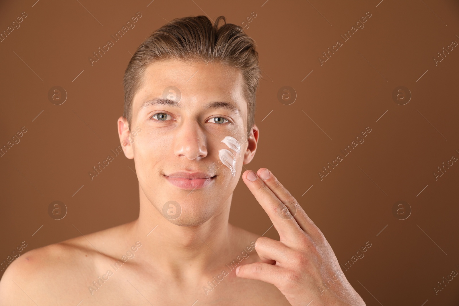 Photo of Handsome young man applying cream onto his face on brown background, space for text