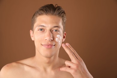 Photo of Handsome young man applying cream onto his face on brown background, space for text