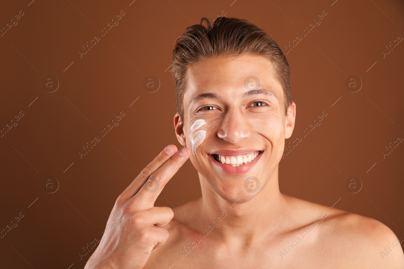 Photo of Handsome young man applying cream onto his face on brown background, space for text