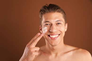 Photo of Handsome young man applying cream onto his face on brown background, space for text