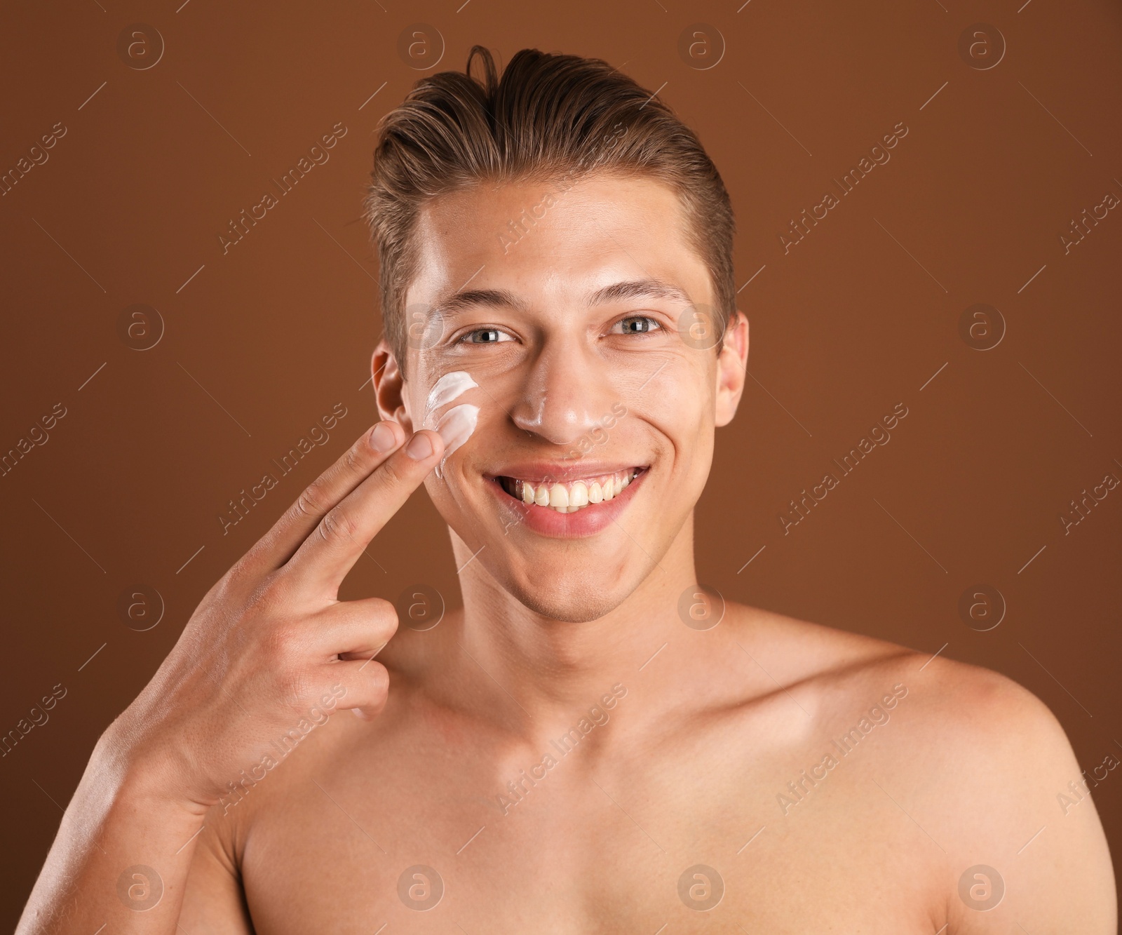 Photo of Handsome young man applying cream onto his face on brown background