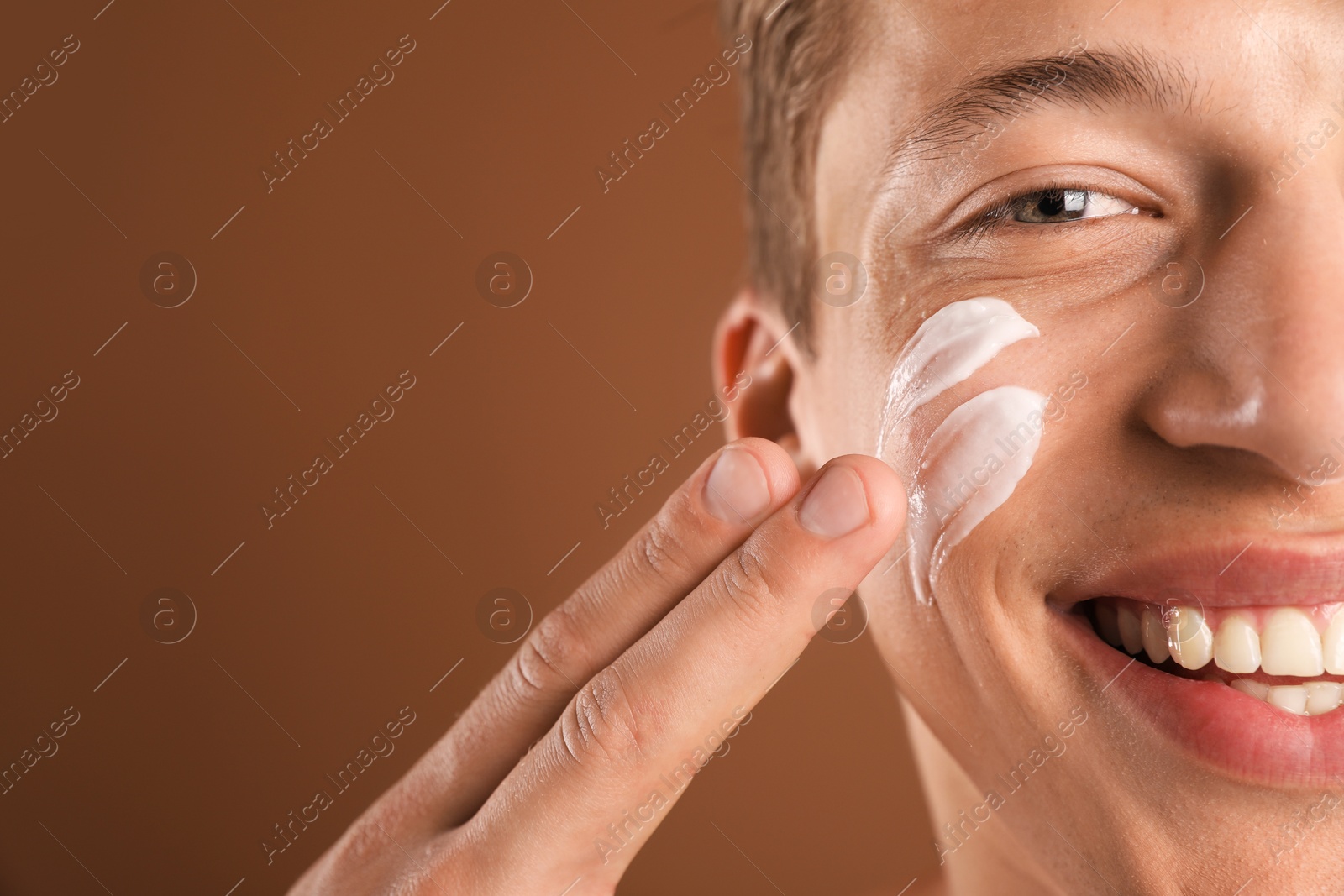 Photo of Handsome young man applying cream onto his face on brown background, closeup. Space for text