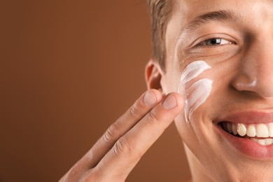 Photo of Handsome young man applying cream onto his face on brown background, closeup. Space for text