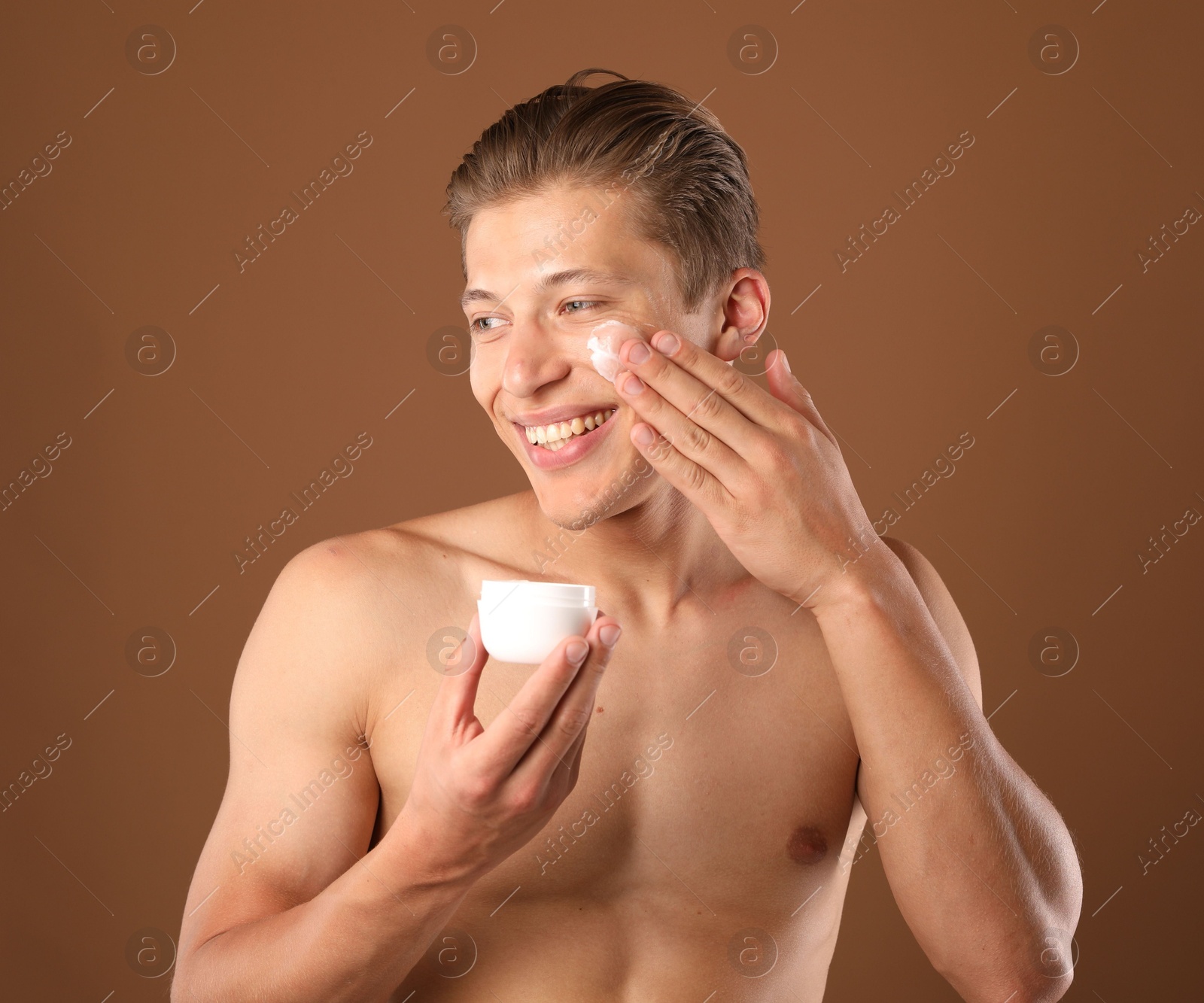Photo of Handsome young man applying cream onto his face on brown background