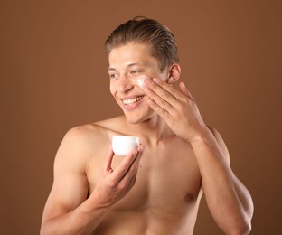 Photo of Handsome young man applying cream onto his face on brown background