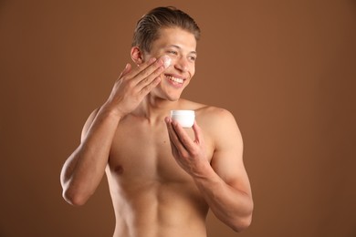 Photo of Handsome young man applying cream onto his face on brown background