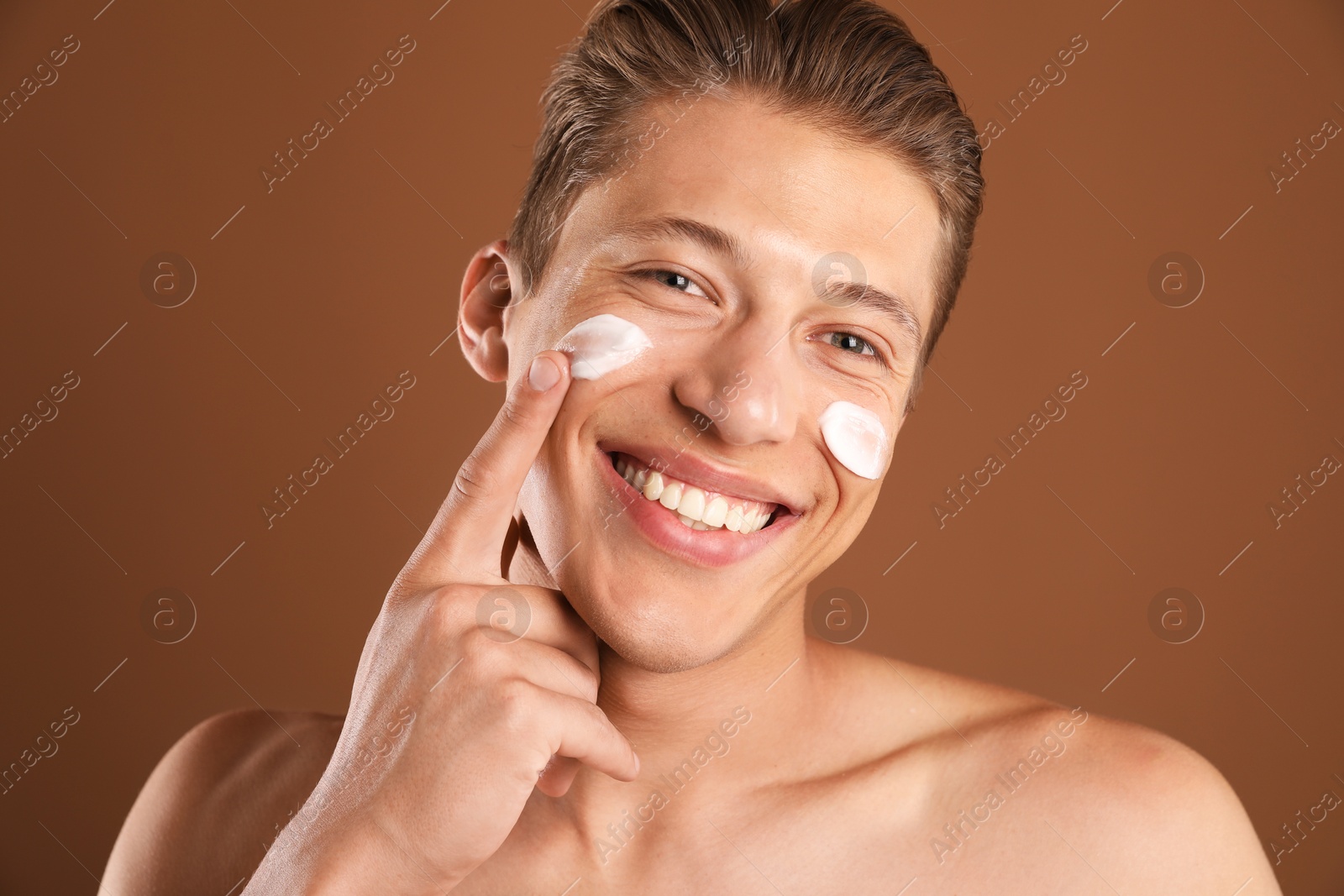 Photo of Handsome young man applying cream onto his face on brown background