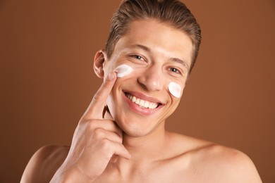 Photo of Handsome young man applying cream onto his face on brown background