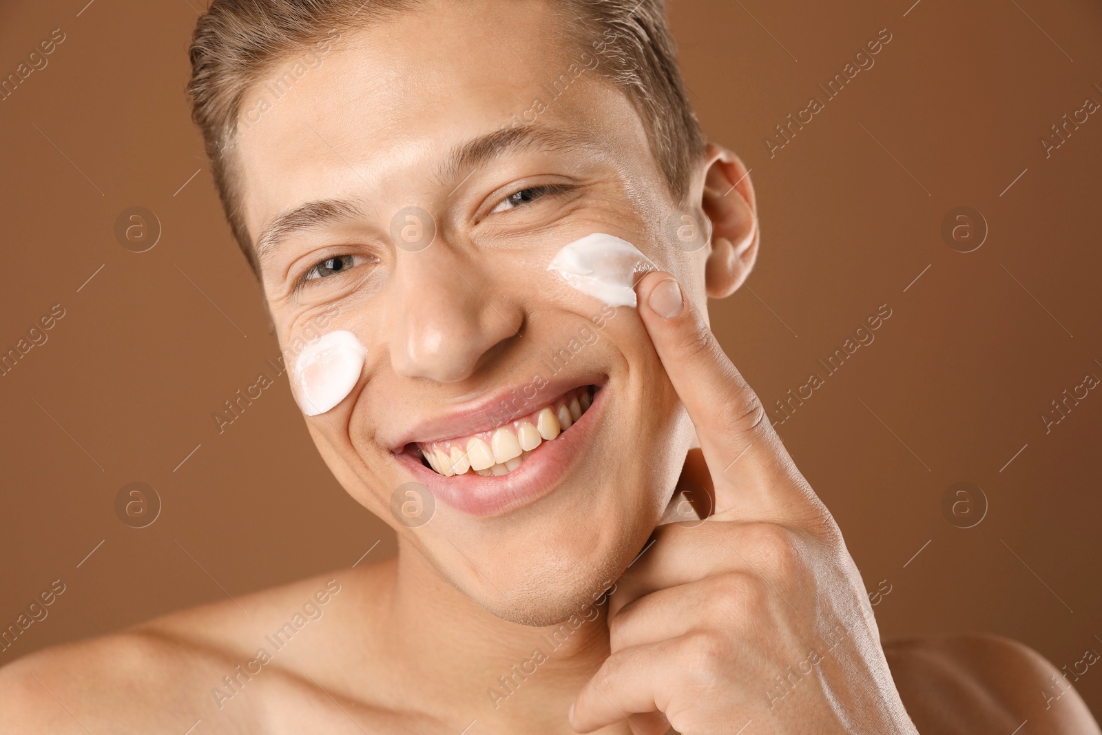 Photo of Handsome young man applying cream onto his face on brown background, closeup