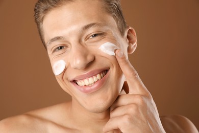 Photo of Handsome young man applying cream onto his face on brown background, closeup