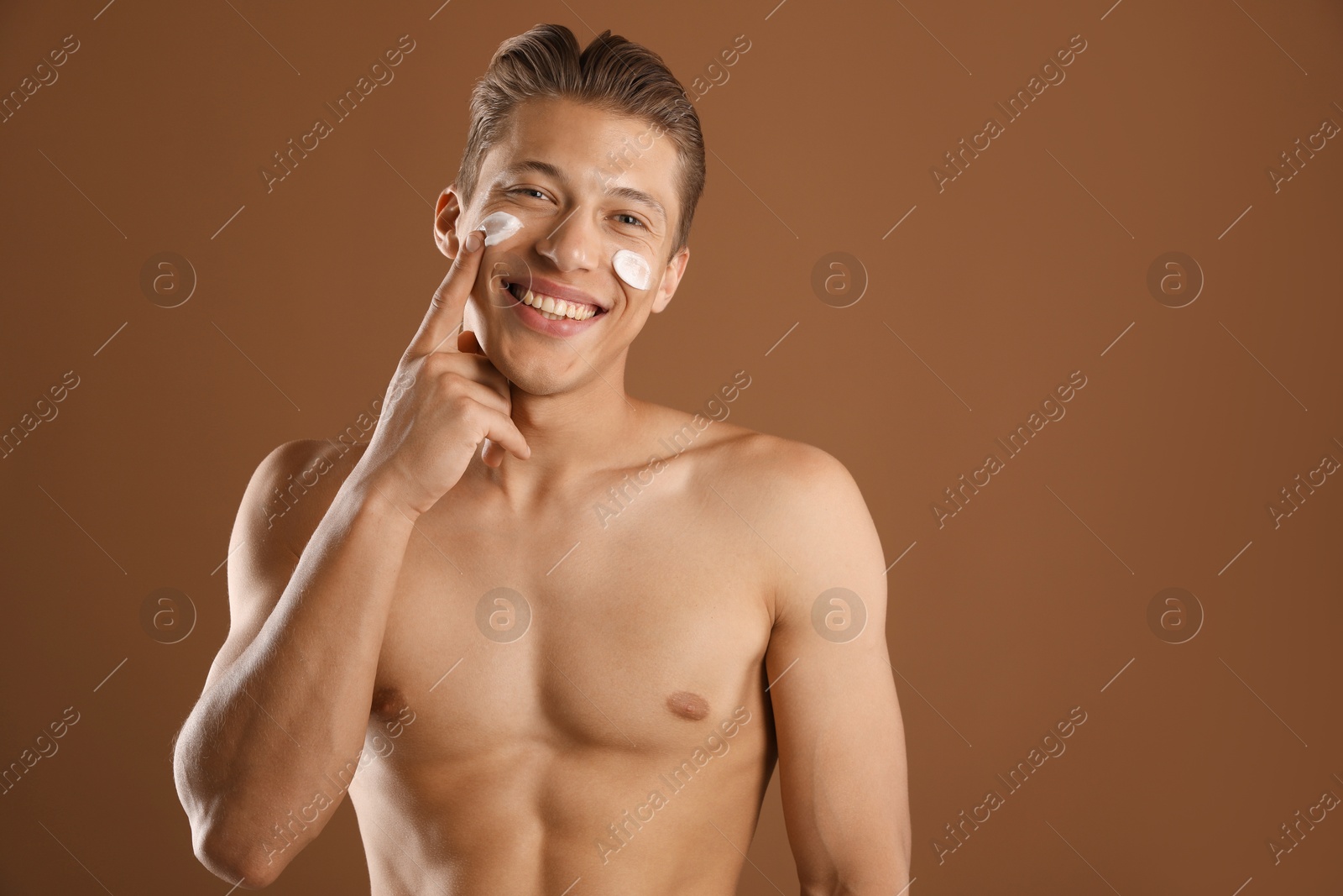 Photo of Handsome young man applying cream onto his face on brown background, space for text