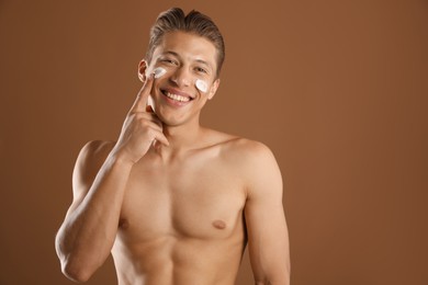 Photo of Handsome young man applying cream onto his face on brown background, space for text