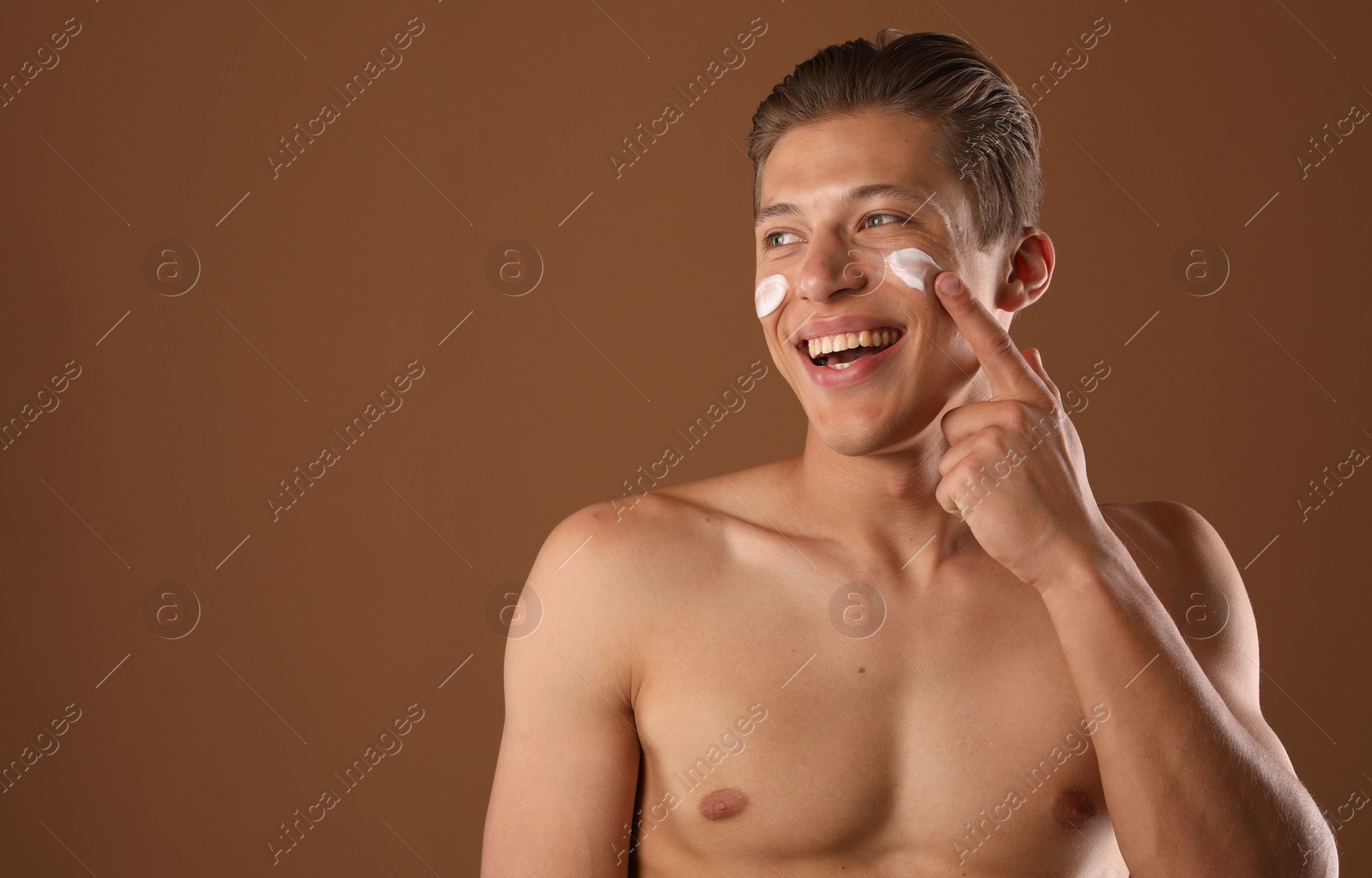 Photo of Handsome young man applying cream onto his face on brown background, space for text