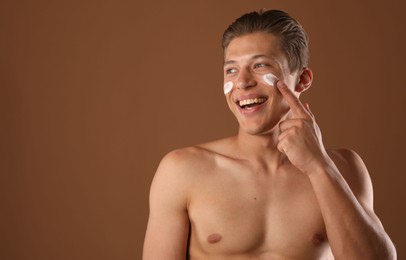 Photo of Handsome young man applying cream onto his face on brown background, space for text