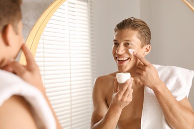 Photo of Handsome young man applying cream onto his face near mirror in bathroom