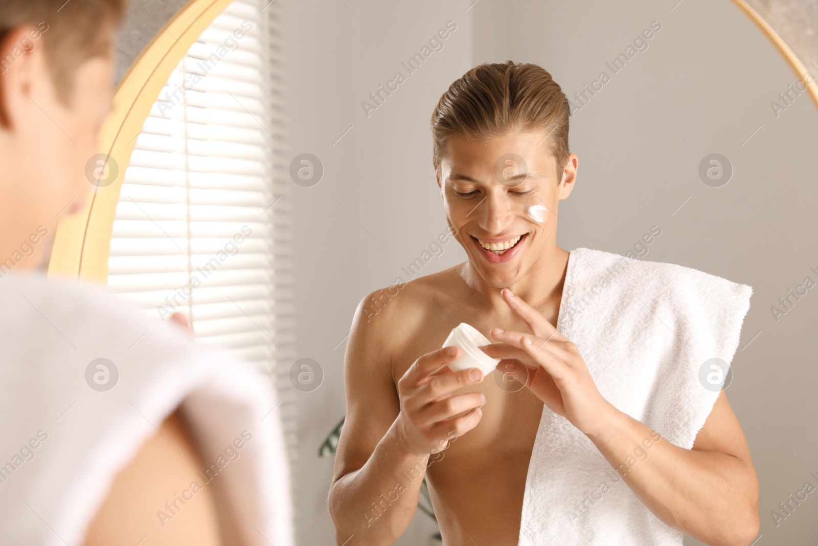 Photo of Handsome young man with jar of face cream near mirror in bathroom