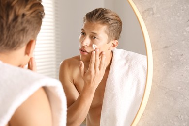 Photo of Handsome young man applying cream onto his face near mirror in bathroom
