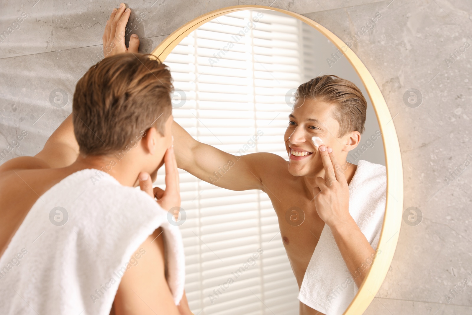 Photo of Handsome young man applying cream onto his face near mirror in bathroom