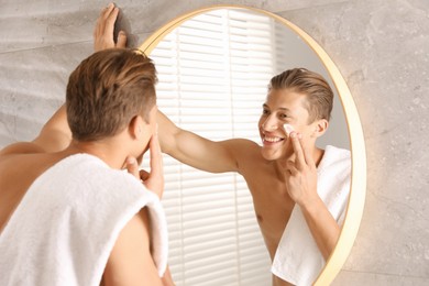 Photo of Handsome young man applying cream onto his face near mirror in bathroom