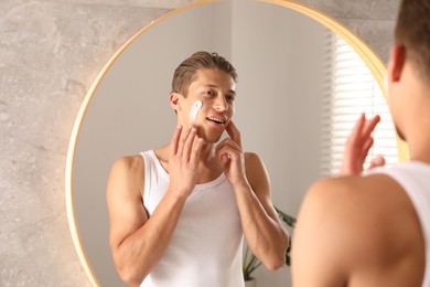 Photo of Handsome young man applying cream onto his face near mirror in bathroom