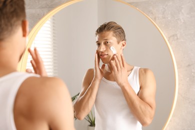 Photo of Handsome young man applying cream onto his face near mirror in bathroom