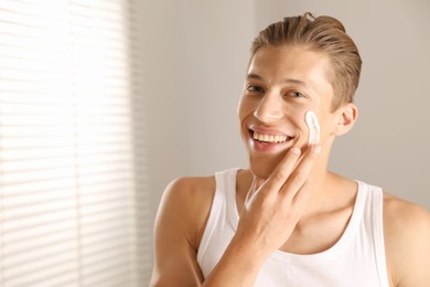 Photo of Handsome young man applying cream onto his face indoors, space for text