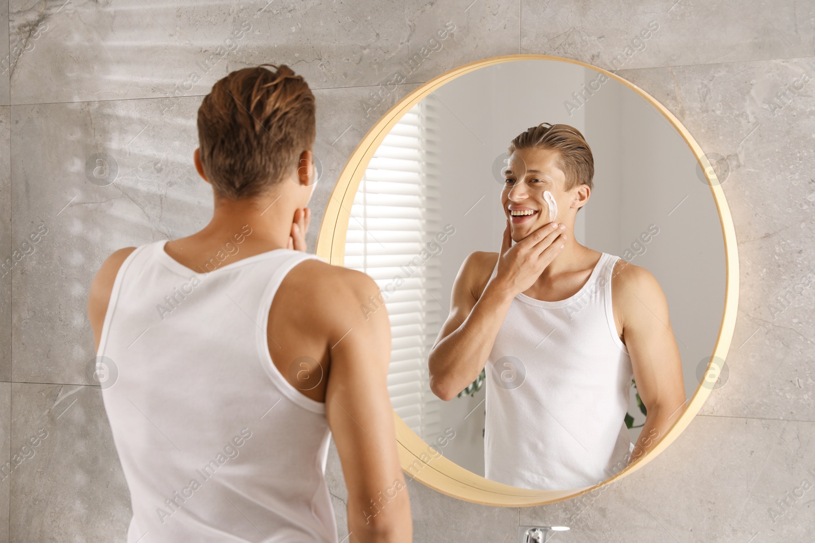 Photo of Handsome young man applying cream onto his face near mirror in bathroom