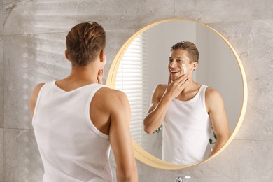 Photo of Handsome young man applying cream onto his face near mirror in bathroom