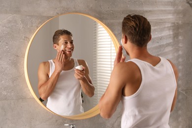 Photo of Handsome young man applying cream onto his face near mirror in bathroom