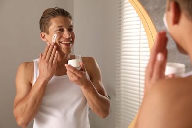 Photo of Handsome young man applying cream onto his face near mirror in bathroom