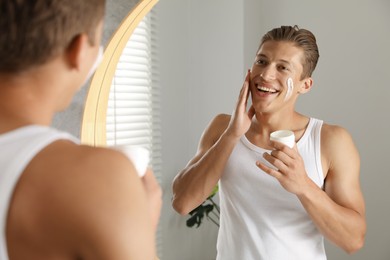 Photo of Handsome young man applying cream onto his face near mirror in bathroom