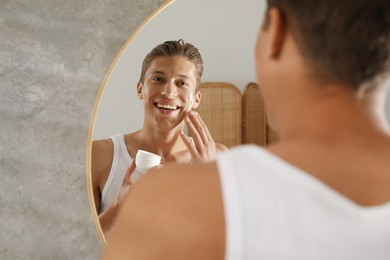 Photo of Handsome young man applying cream onto his face near mirror in bathroom
