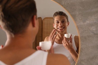 Photo of Handsome young man applying cream onto his face near mirror in bathroom
