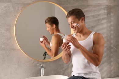 Photo of Handsome young man with jar of face cream near mirror in bathroom