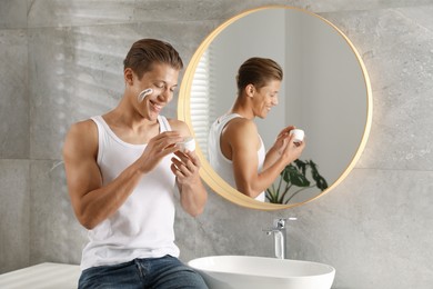 Photo of Handsome young man with jar of face cream near mirror in bathroom