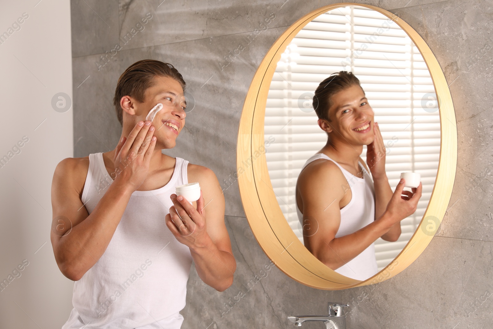 Photo of Handsome young man applying cream onto his face near mirror in bathroom