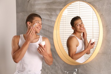 Photo of Handsome young man applying cream onto his face near mirror in bathroom