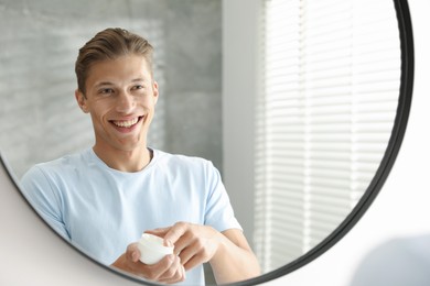 Photo of Handsome young man with jar of face cream near mirror at home