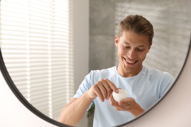 Photo of Handsome young man with jar of face cream near mirror at home