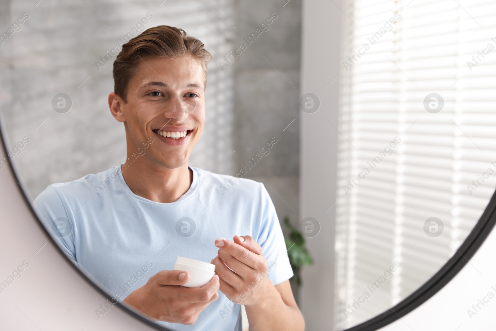 Photo of Handsome young man with jar of face cream near mirror at home