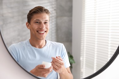 Photo of Handsome young man with jar of face cream near mirror at home