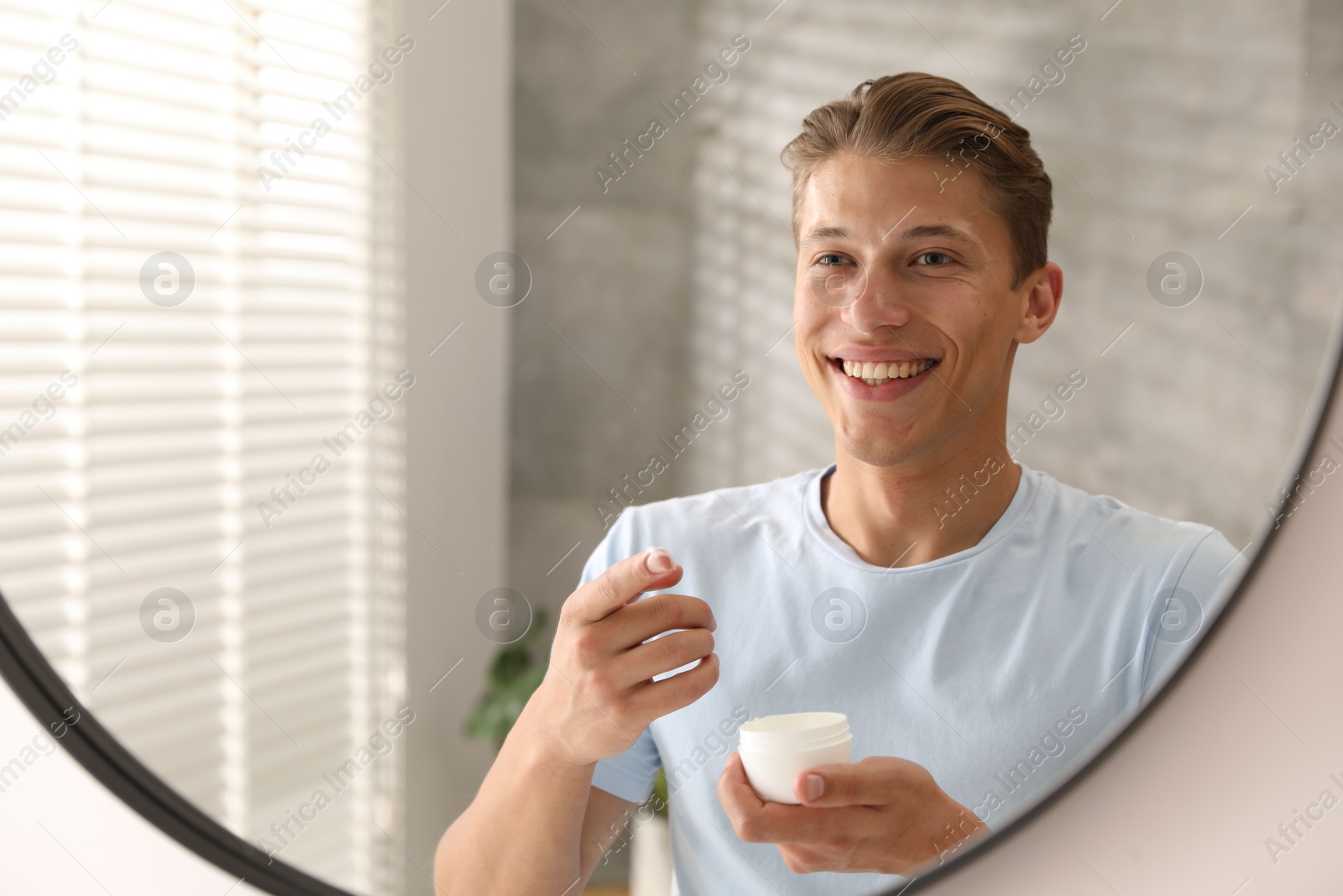 Photo of Handsome young man with jar of face cream near mirror at home