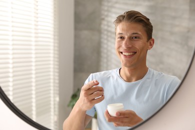 Photo of Handsome young man with jar of face cream near mirror at home