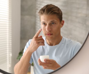 Photo of Handsome young man applying cream onto his face near mirror at home