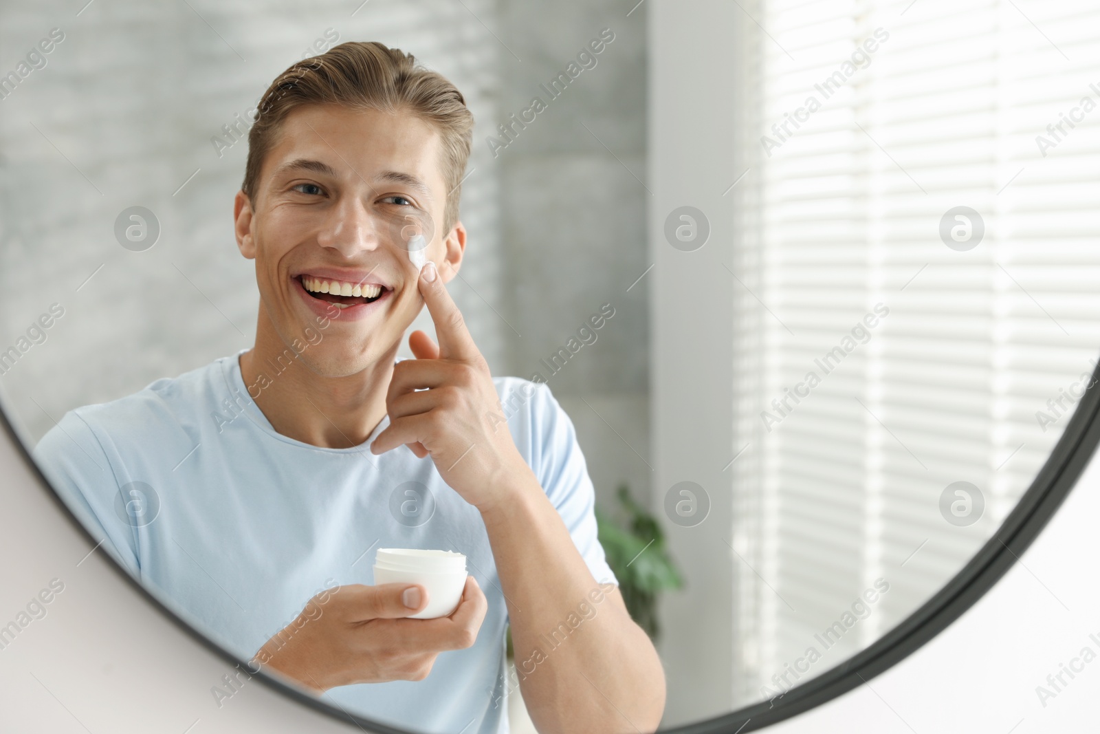 Photo of Handsome young man applying cream onto his face near mirror at home